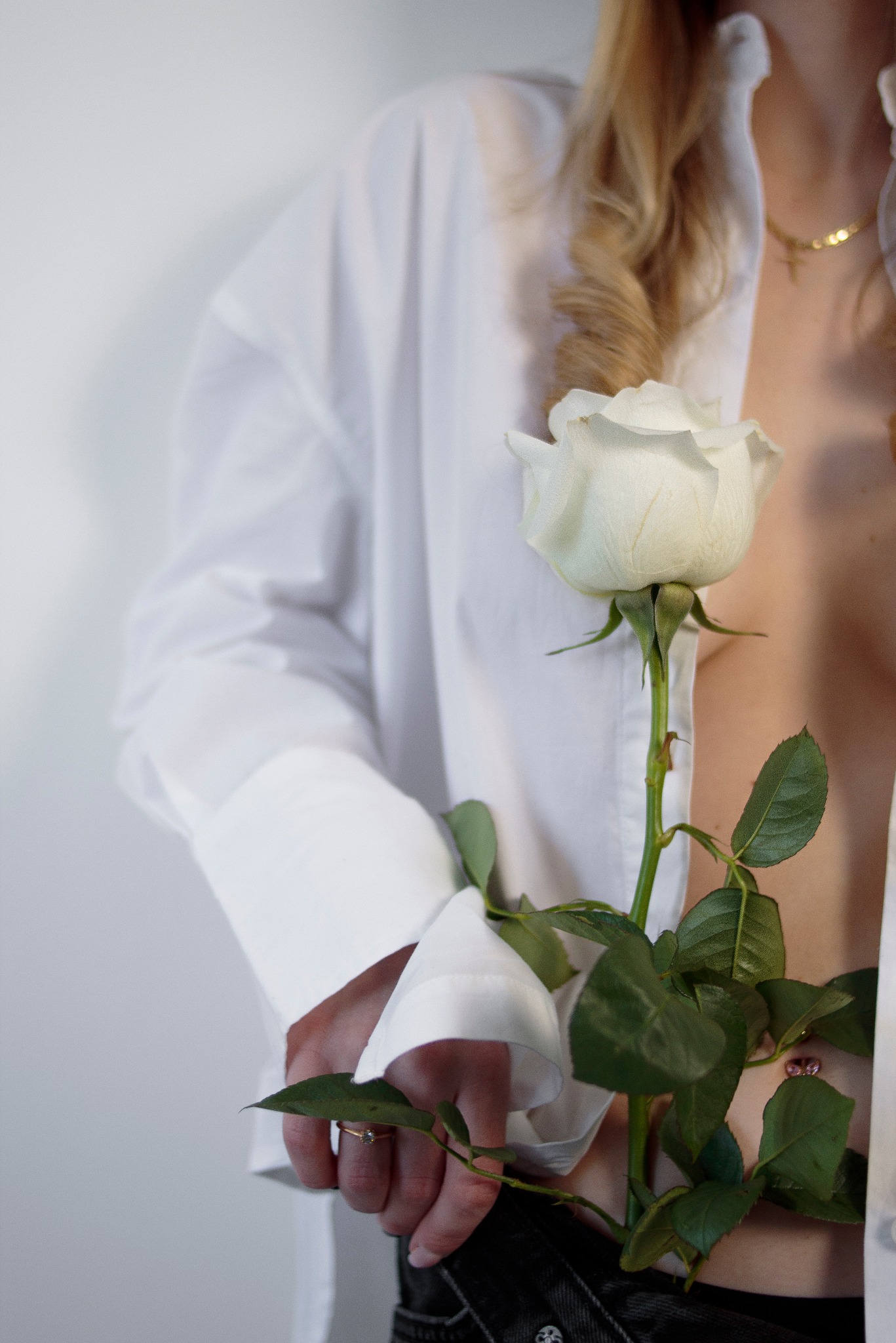 Kobieca sesja: blondynka w białej koszuli, z delikatnym złotym łańcuszkiem, trzyma białą różę na tle jasnej ściany. Widoczne dłonie z pierścionkiem i fragment czarnych spodni.