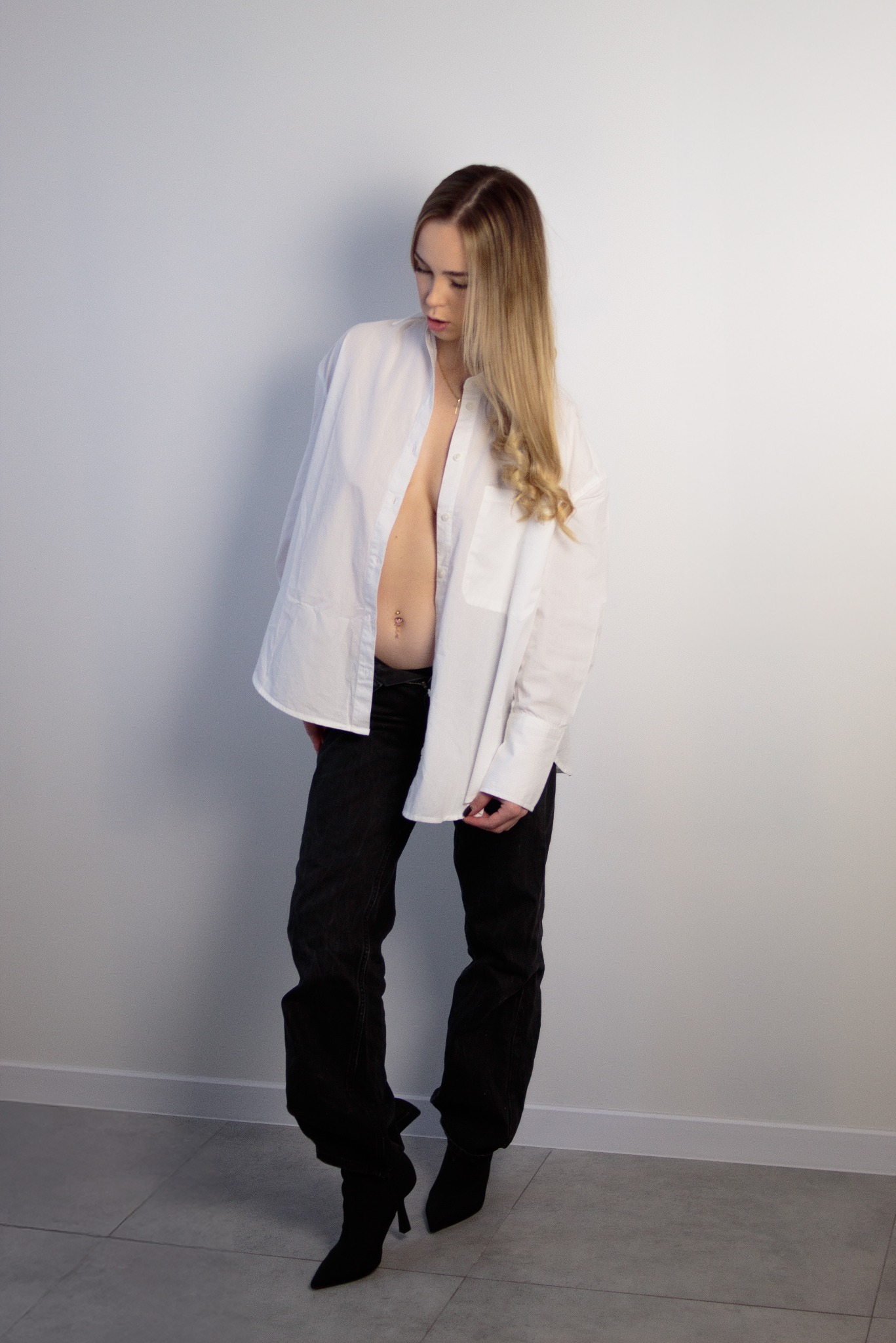 Blondwłosa kobieta w rozpiętej białej koszuli i czarnych dżinsach pozuje na tle białej ściany. Widoczny piercing w pępku i fragment podłogi z szarych płytek.