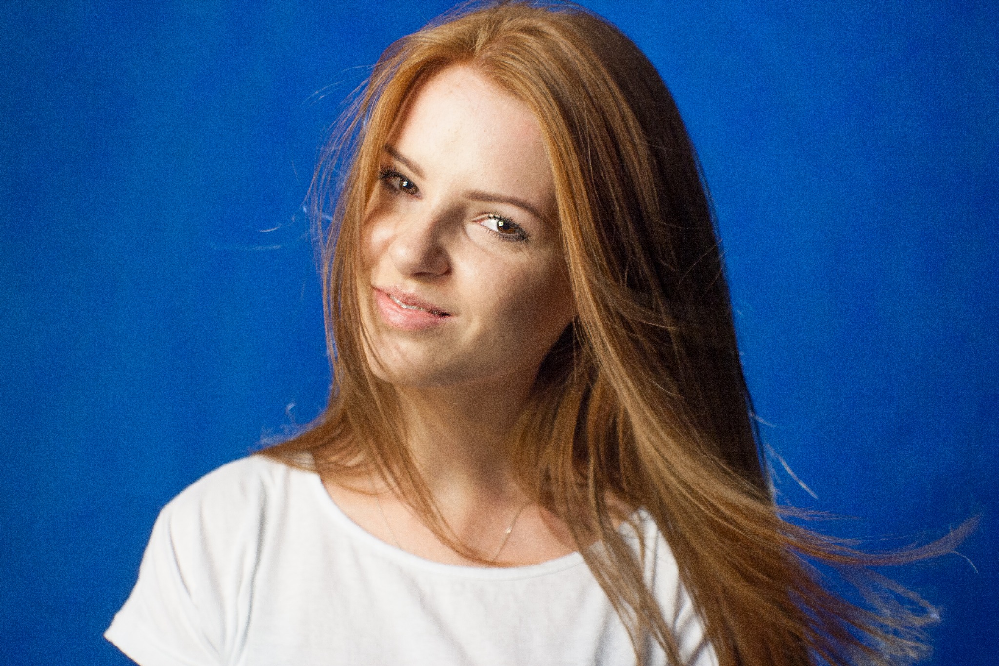 Portret uśmiechniętej, młodej kobiety z długimi, rudymi włosami na tle intensywnego, niebieskiego tła. Ubrana w białą koszulkę, patrzy w obiektyw z lekkim przekrzywieniem głowy.