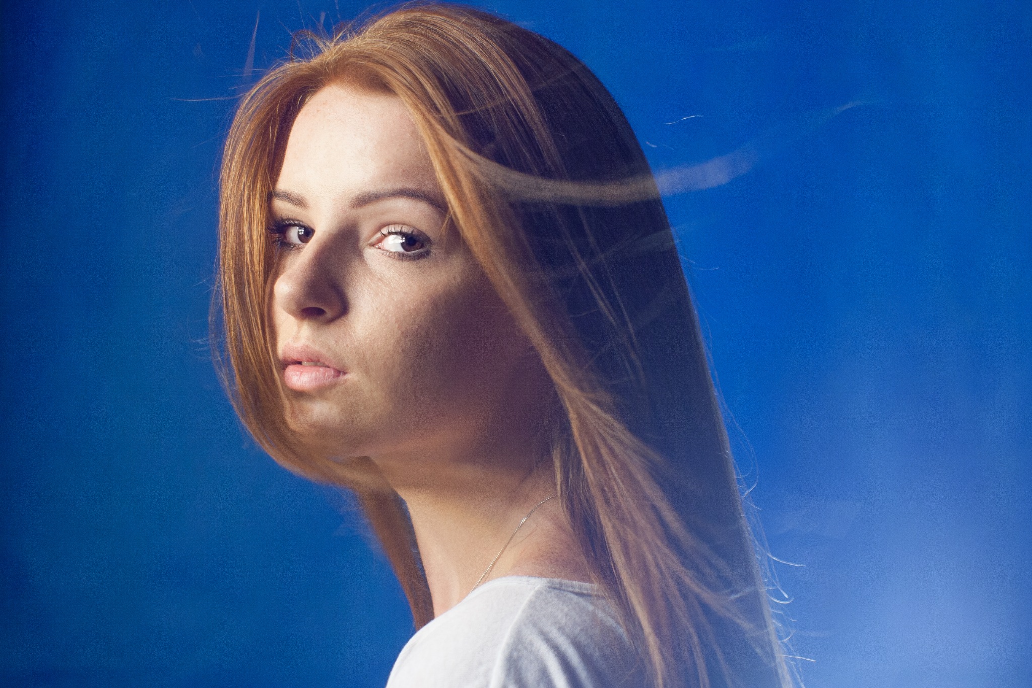 Portret kobiety o rudych włosach, spoglądającej przez ramię na tle jednolitego, niebieskiego tła. Delikatny makijaż, subtelna biżuteria. Włosy rozwiane przez wiatr.