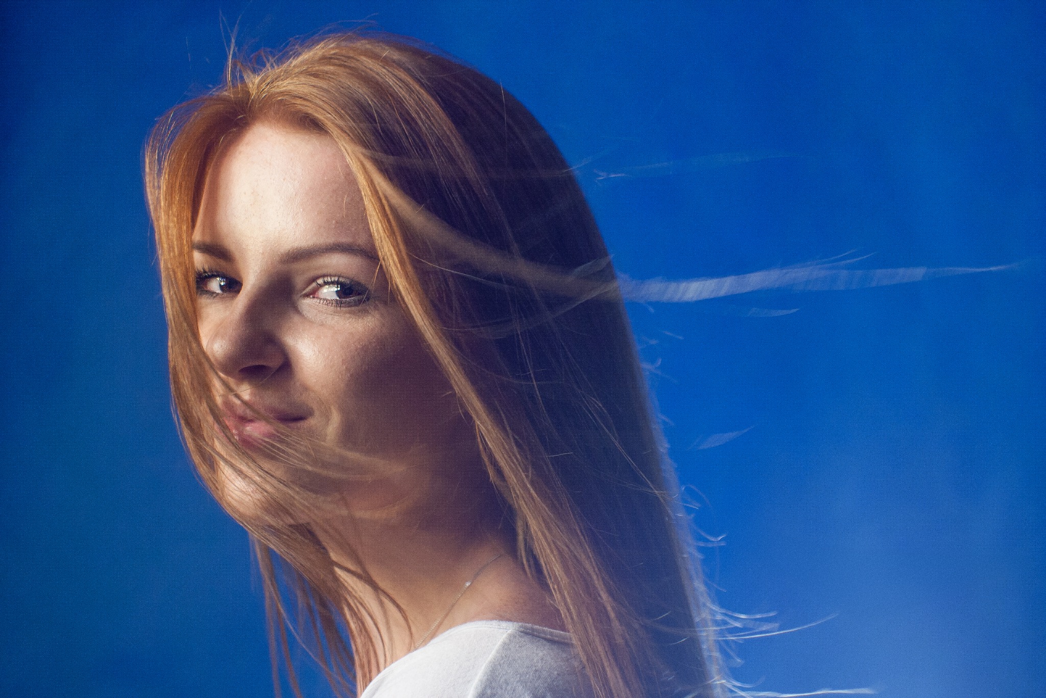 Portret kobiety o rudych włosach, spoglądającej przez ramię na tle intensywnego, błękitnego tła. Włosy delikatnie rozwiane, podkreślają naturalność ujęcia.