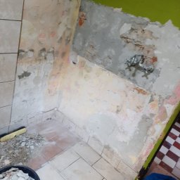 Daniel Gruszczyński - Demontaż płytek w rogu łazienki, widoczne fragmenty starej instalacji wodnej, poziomica i wiadro z gruzem. Przygotowanie do remontu, widok z góry.