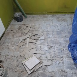 Daniel Gruszczyński - Demontaż starej podłogi z płytek. Fragmenty płytek na podłożu, widoczne ślady po kleju. Wiaderka z odłamkami i narzędzia w tle. Przygotowanie do remontu.