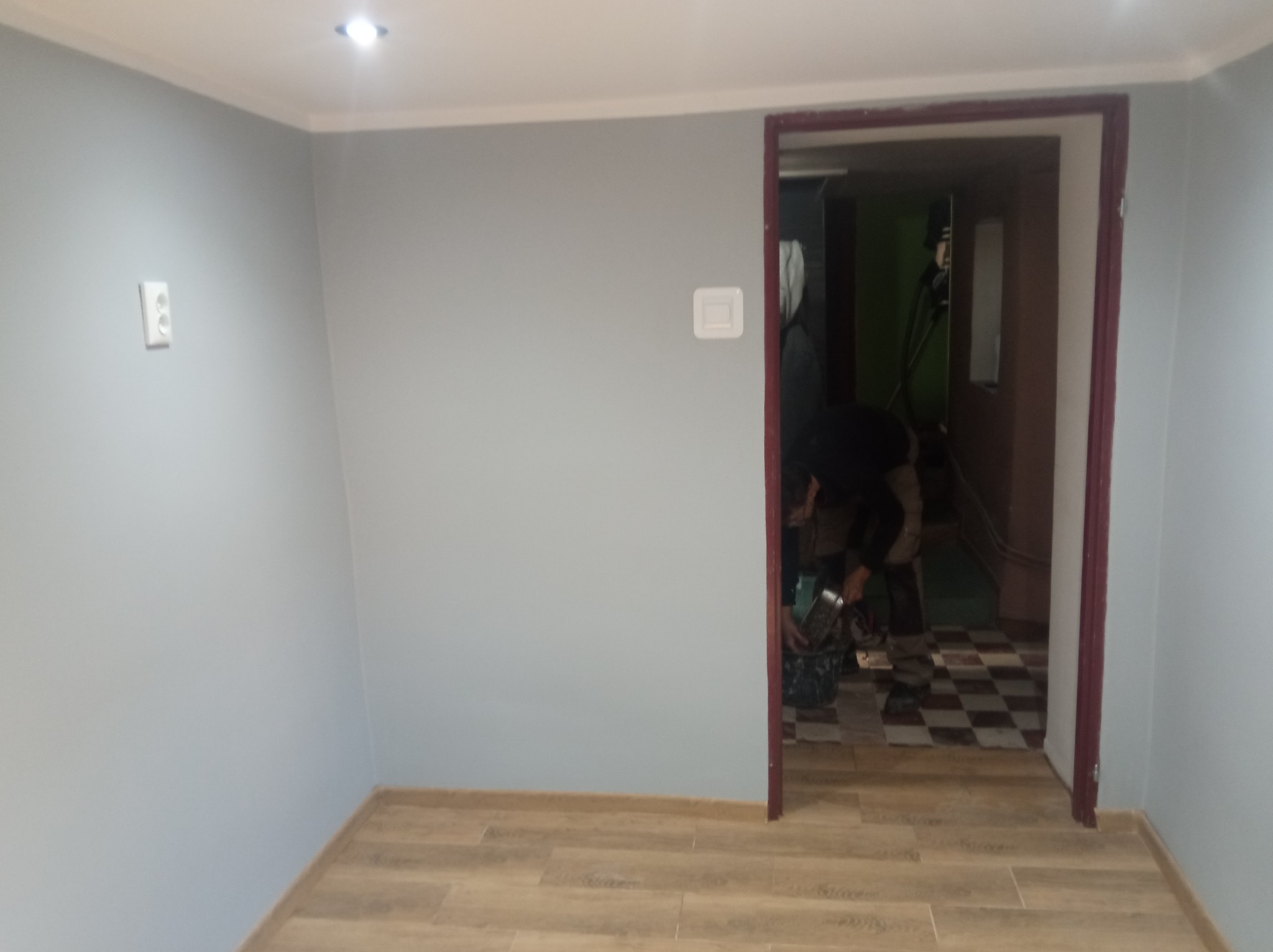 Jasnoszary pokój po malowaniu z drewnopodobną podłogą. W tle widoczne narzędzia i osoba pracująca w pomieszczeniu z szachownicą na podłodze.