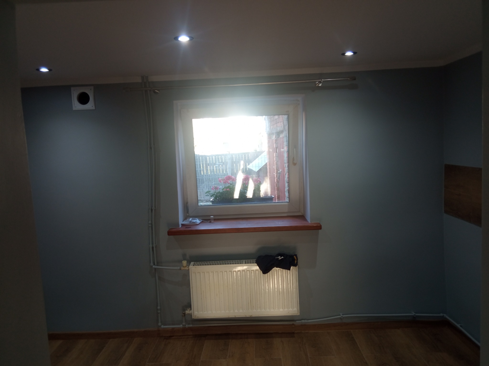 Pokój po remoncie z szarymi ścianami, drewnianą podłogą i białym grzejnikiem pod oknem. Widoczne rury grzewcze i punktowe oświetlenie sufitowe.