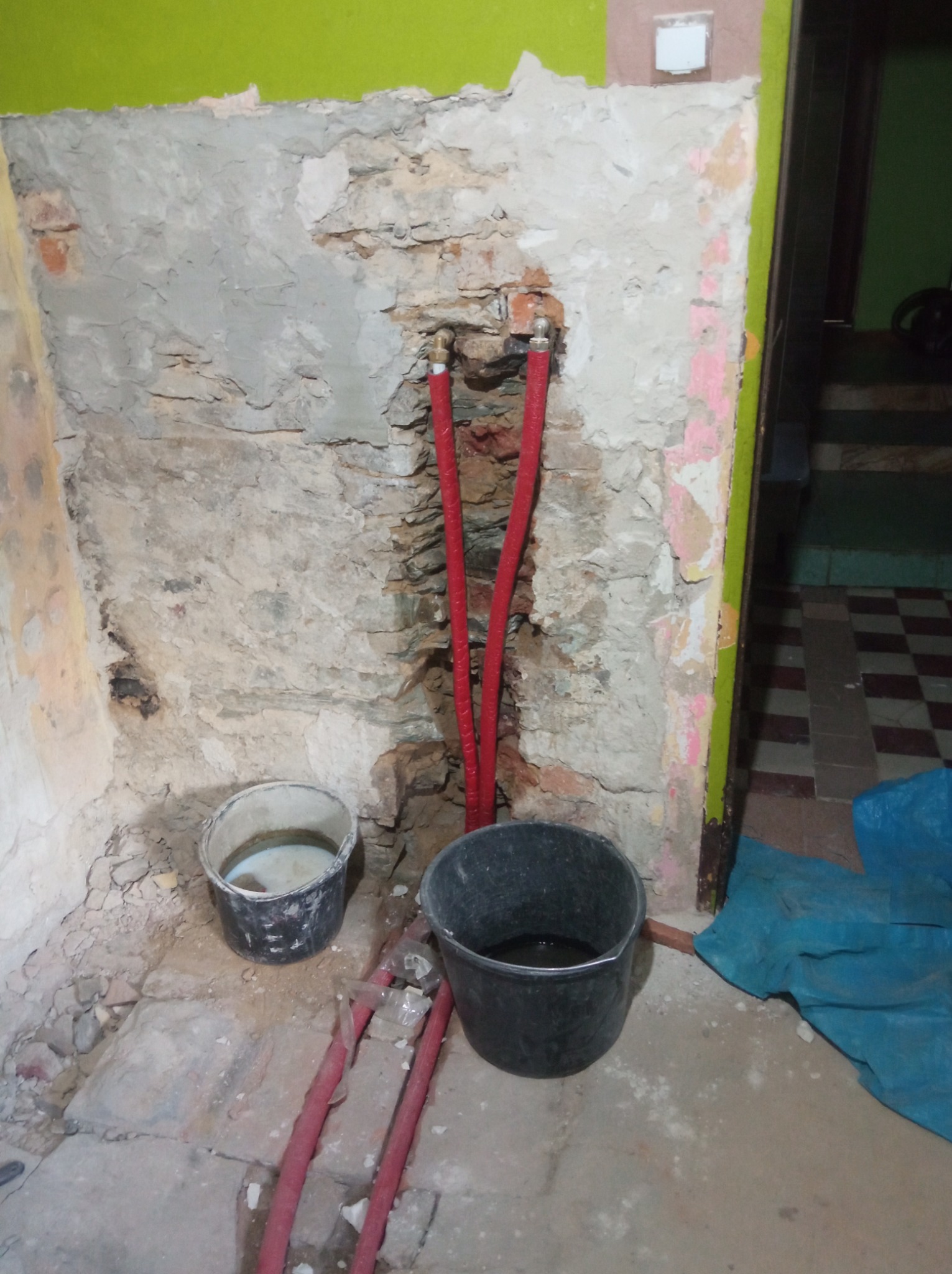 Fragment ściany z odsłoniętą instalacją hydrauliczną, dwa wiadra na podłodze. Widoczne ślady po kuciu i przygotowaniu do dalszych prac wykończeniowych.