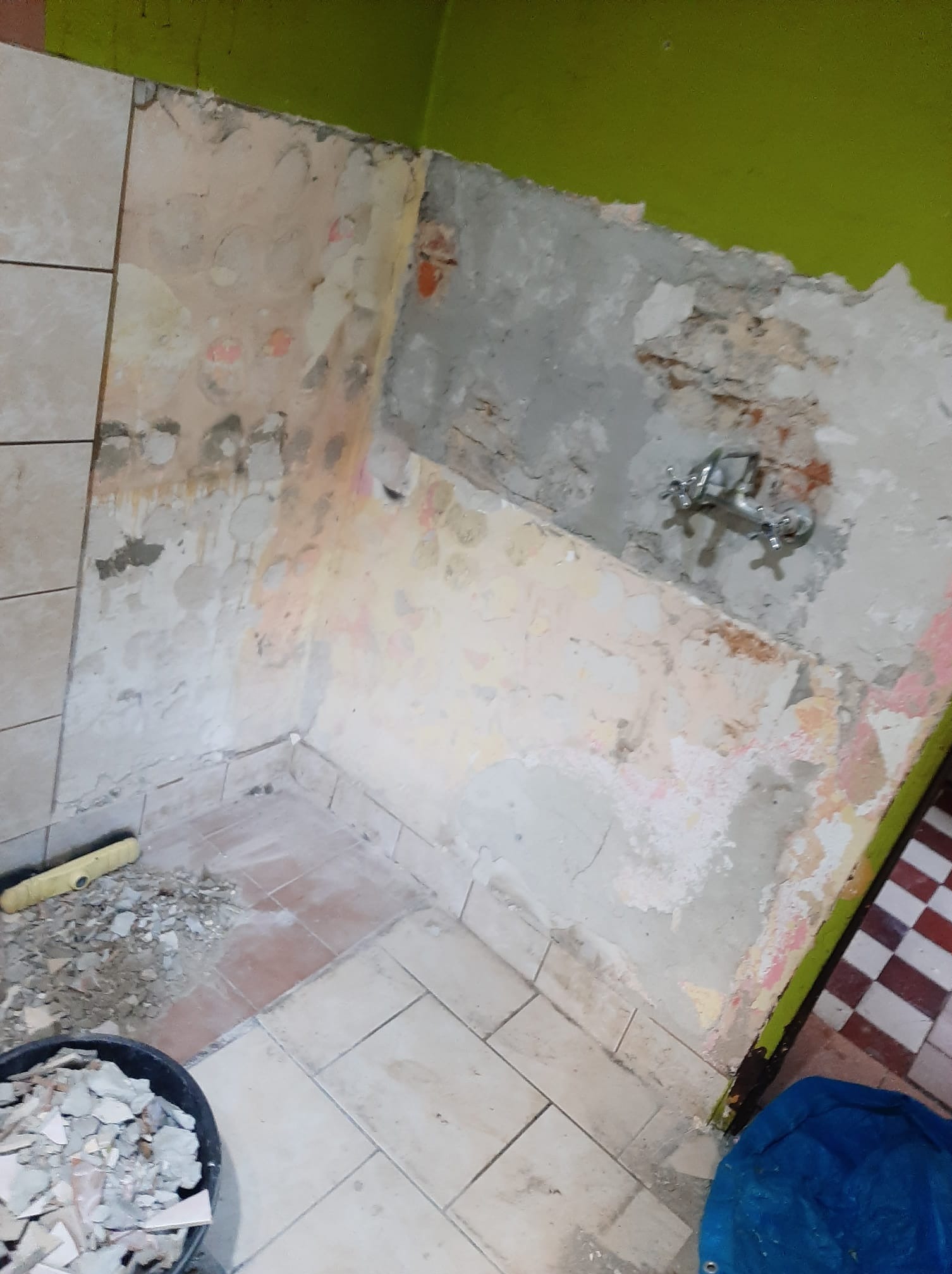 Demontaż płytek w rogu łazienki, widoczne fragmenty starej instalacji wodnej, poziomica i wiadro z gruzem. Przygotowanie do remontu, widok z góry.