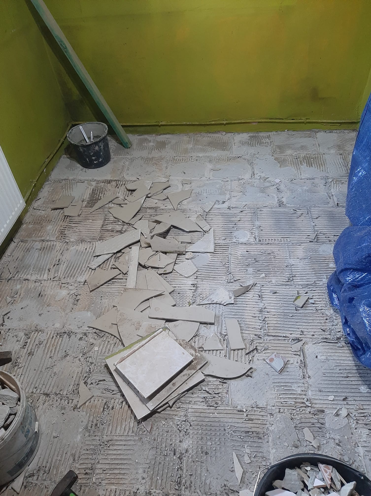 Demontaż starej podłogi z płytek. Fragmenty płytek na podłożu, widoczne ślady po kleju. Wiaderka z odłamkami i narzędzia w tle. Przygotowanie do remontu.
