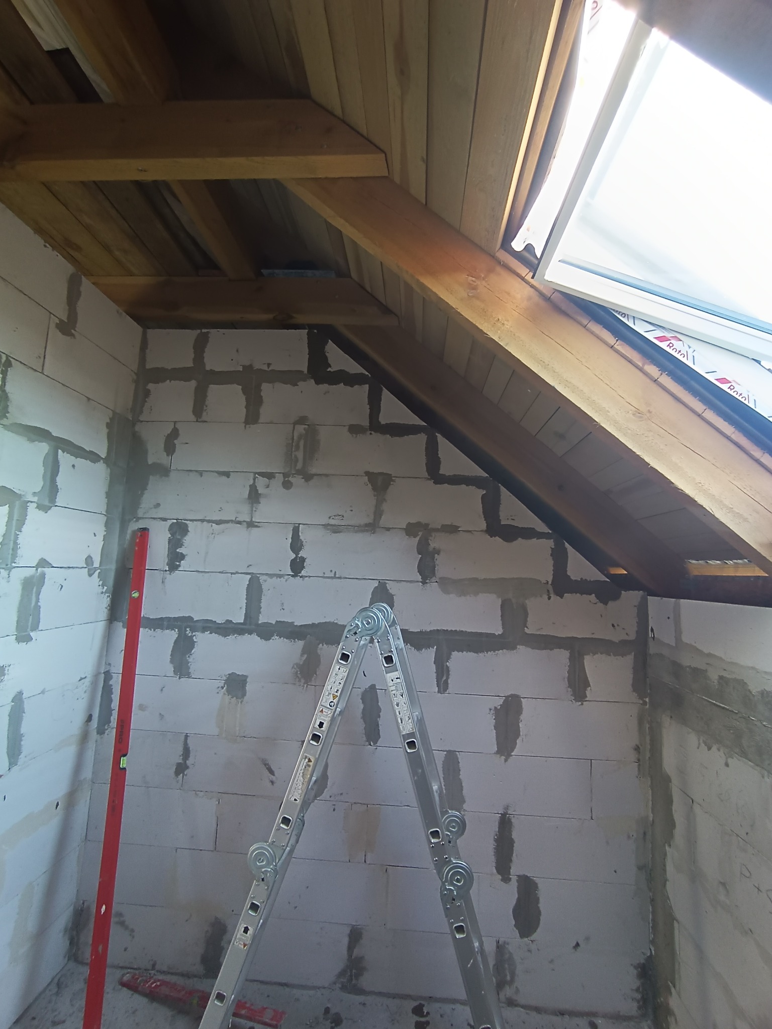 Surowe wnętrze poddasza z oknem dachowym, widoczne bloczki, drewniana konstrukcja dachu, drabina i poziomica. Etap prac wykończeniowych.
