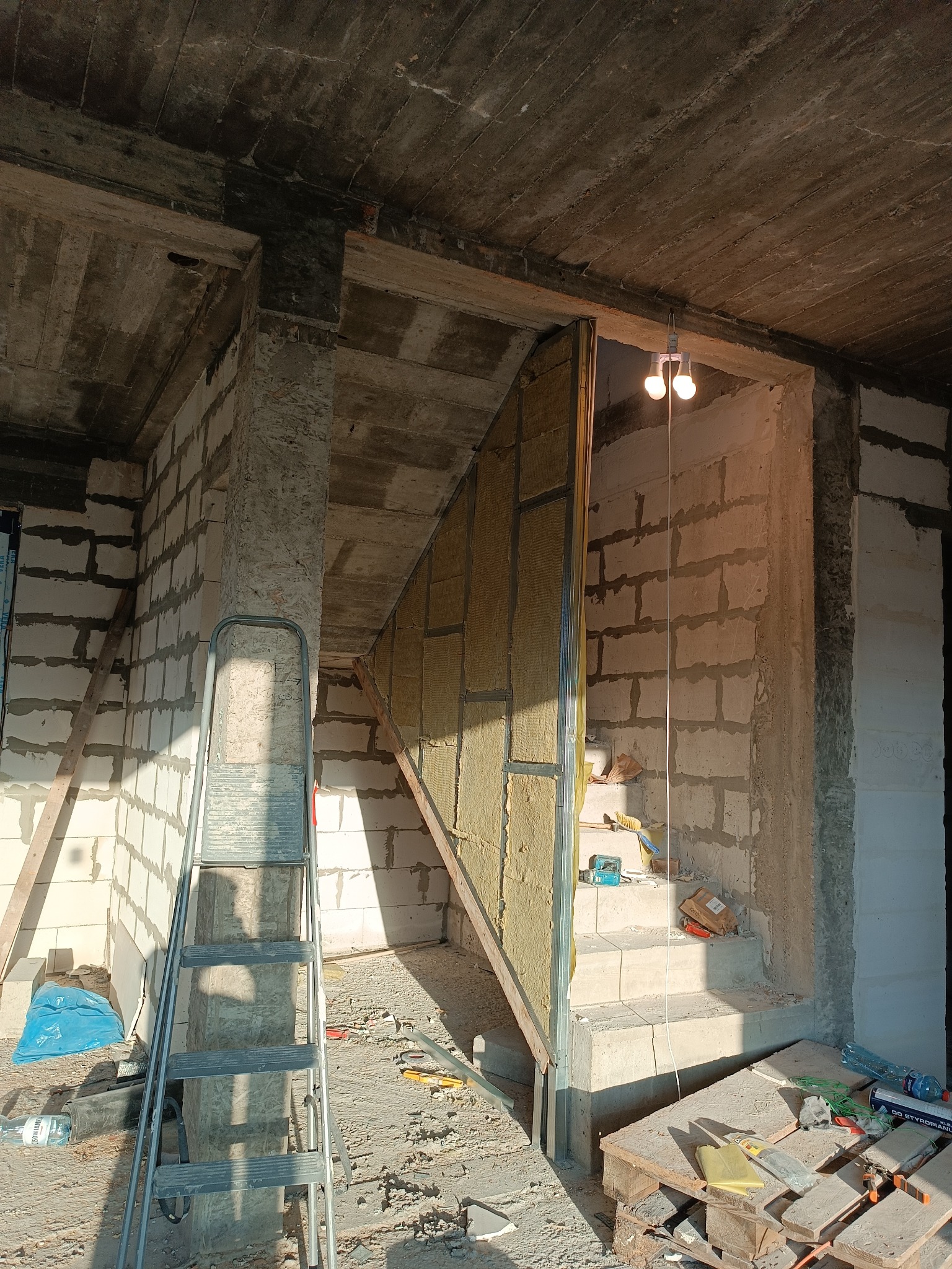 Ocieplenie poddasza wełną mineralną, widoczne metalowe profile, surowy stan wykończenia, drabina oparta o filar, narzędzia budowlane na podłodze.