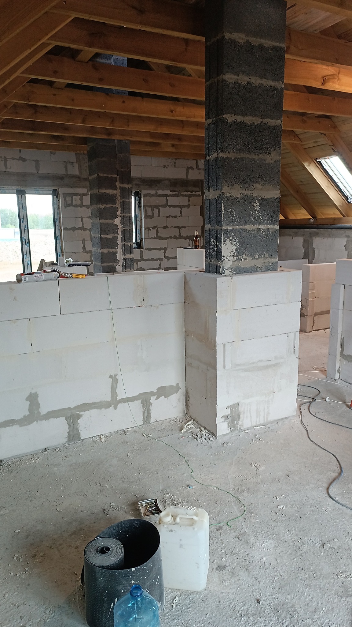 Wnętrze w trakcie budowy: ściany z białych bloczków, szare filary, drewniany strop. Widoczne narzędzia i materiały budowlane. Surowy stan, okna dachowe.