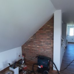 Usługi Remontowo-Budowlane Jacek Baraniewicz - Poddasze w trakcie prac wykończeniowych: fragment ściany z cegły, narzędzia, odkurzacz budowlany na panelach. Widoczne przewody elektryczne.