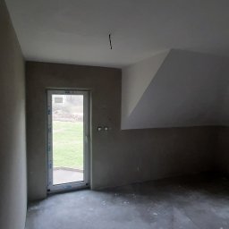 Usługi Remontowo-Budowlane Jacek Baraniewicz - Surowe wnętrze poddasza z oknem balkonowym i widokiem na zieleń. Ściany przygotowane do malowania, sufit biały, podłoga betonowa. Widoczne gniazdka elektryczne.