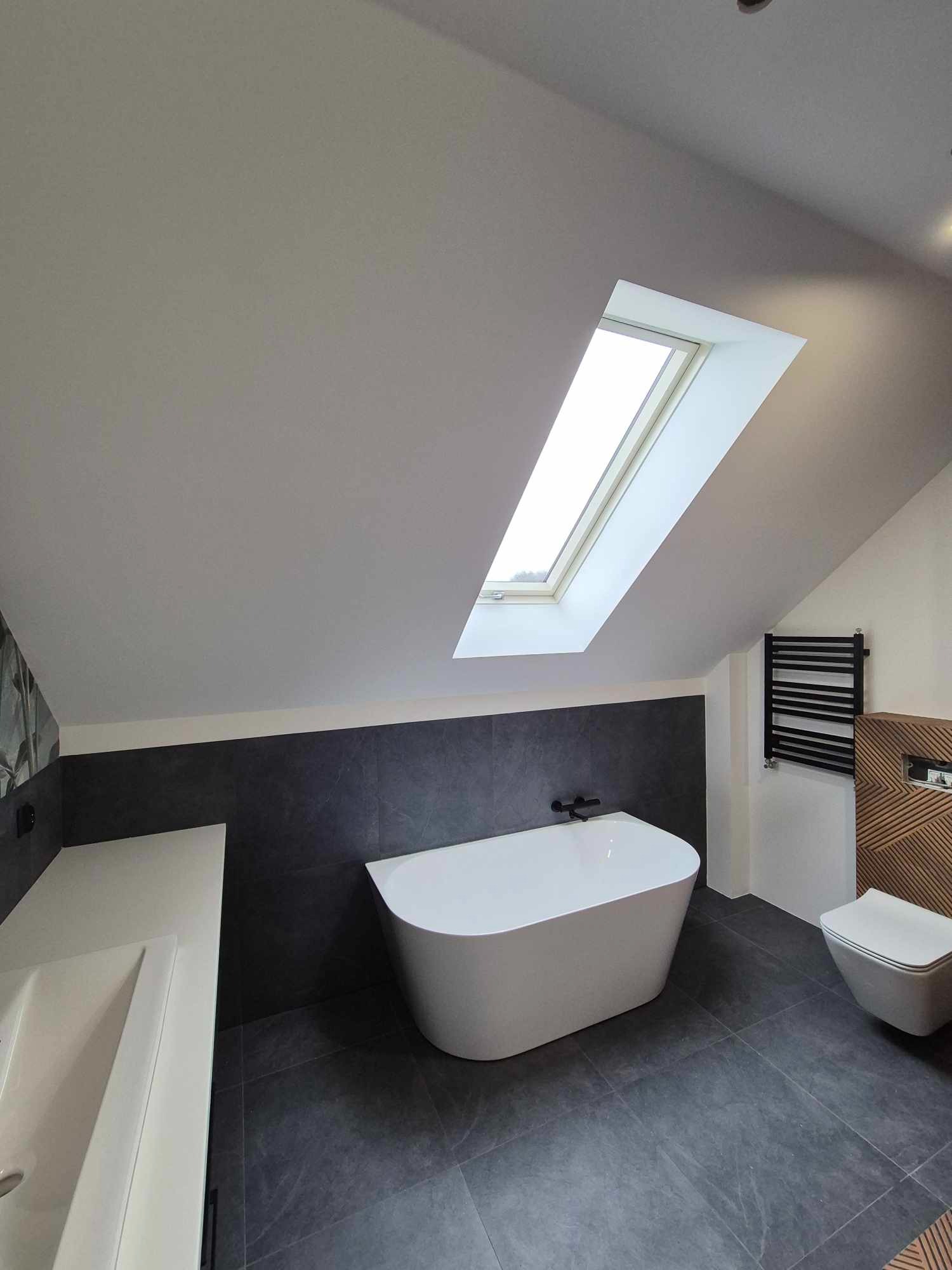 Nowoczesna łazienka na poddaszu z oknem dachowym, wolnostojącą wanną i szarą glazurą. Uwagę zwraca drewniany panel dekoracyjny za toaletą.