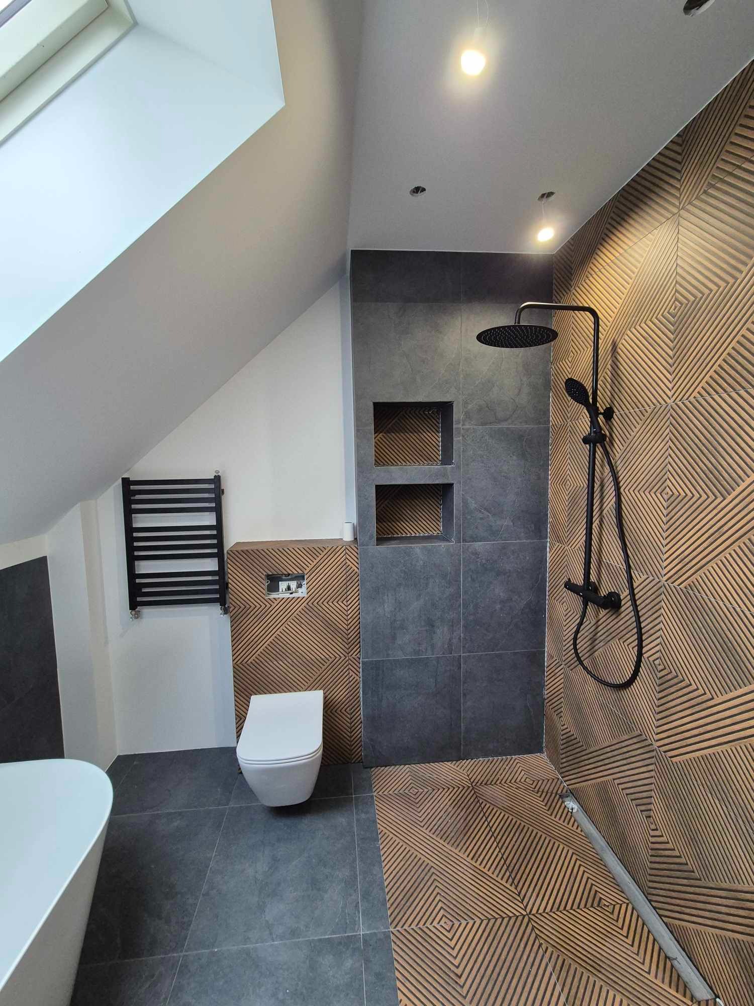 Wykończona łazienka na poddaszu z kabiną prysznicową typu walk-in, toaletą podwieszaną i grzejnikiem drabinkowym. Uwagę zwracają geometryczne płytki na ścianie i podłodze.