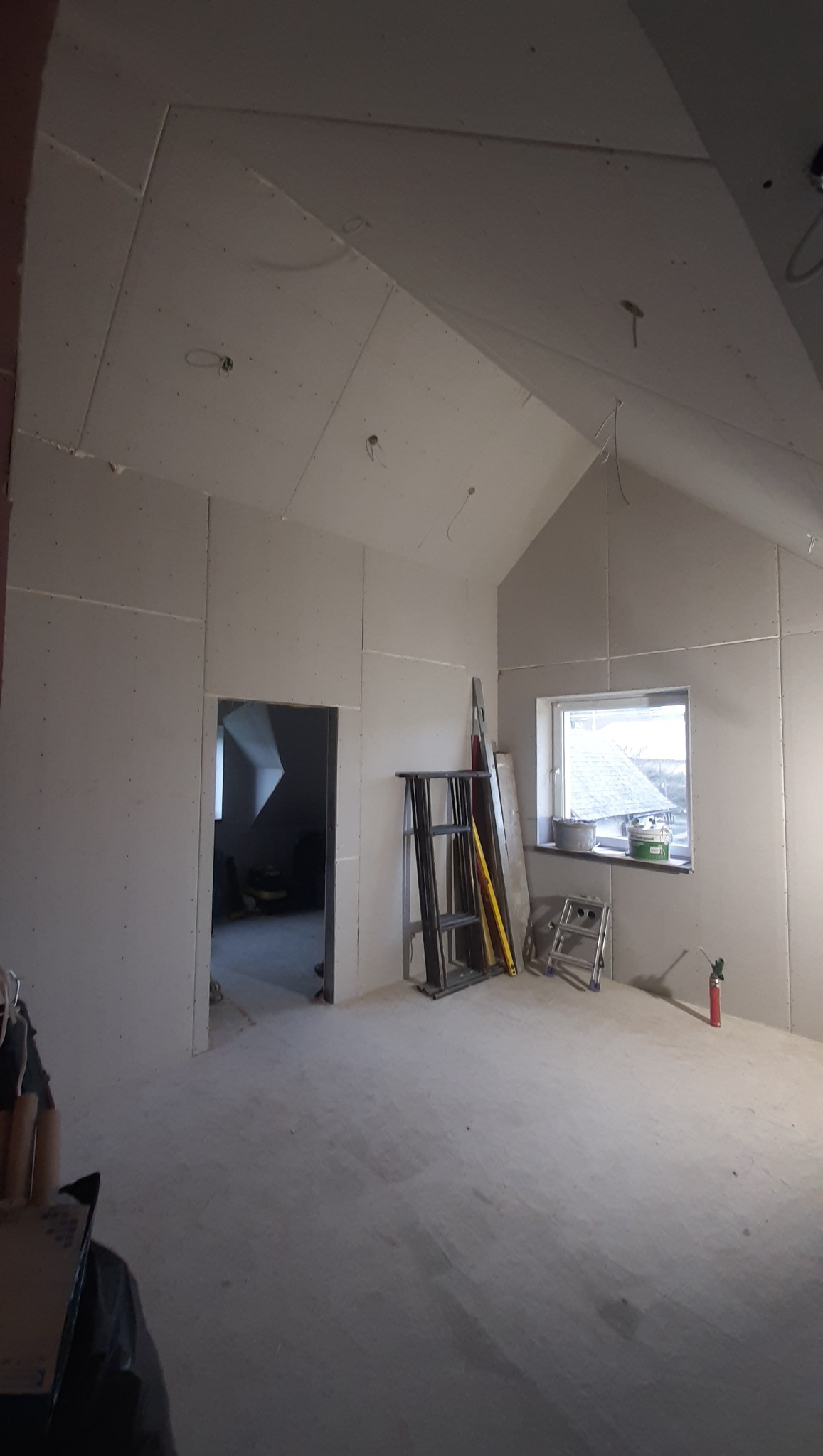 Poddasze w trakcie wykańczania: ściany i sufit obłożone płytami gipsowo-kartonowymi, widoczne drabiny i narzędzia, okno z parapetem. Stan surowy przed malowaniem.