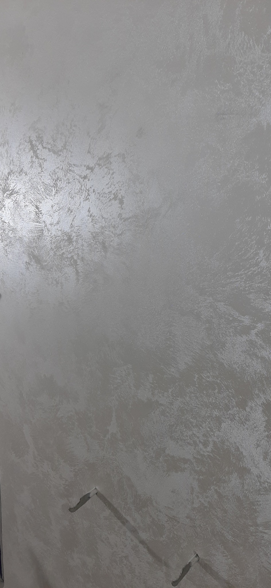 Błyszcząca, szara ściana z dekoracyjną fakturą, oświetlona z lewej strony, z widocznymi elementami instalacji elektrycznej w dolnej części kadru.