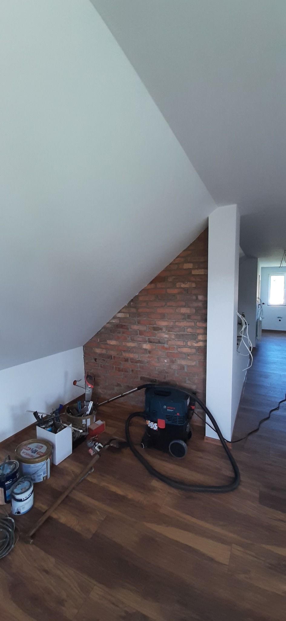 Poddasze w trakcie prac wykończeniowych: fragment ściany z cegły, narzędzia, odkurzacz budowlany na panelach. Widoczne przewody elektryczne.