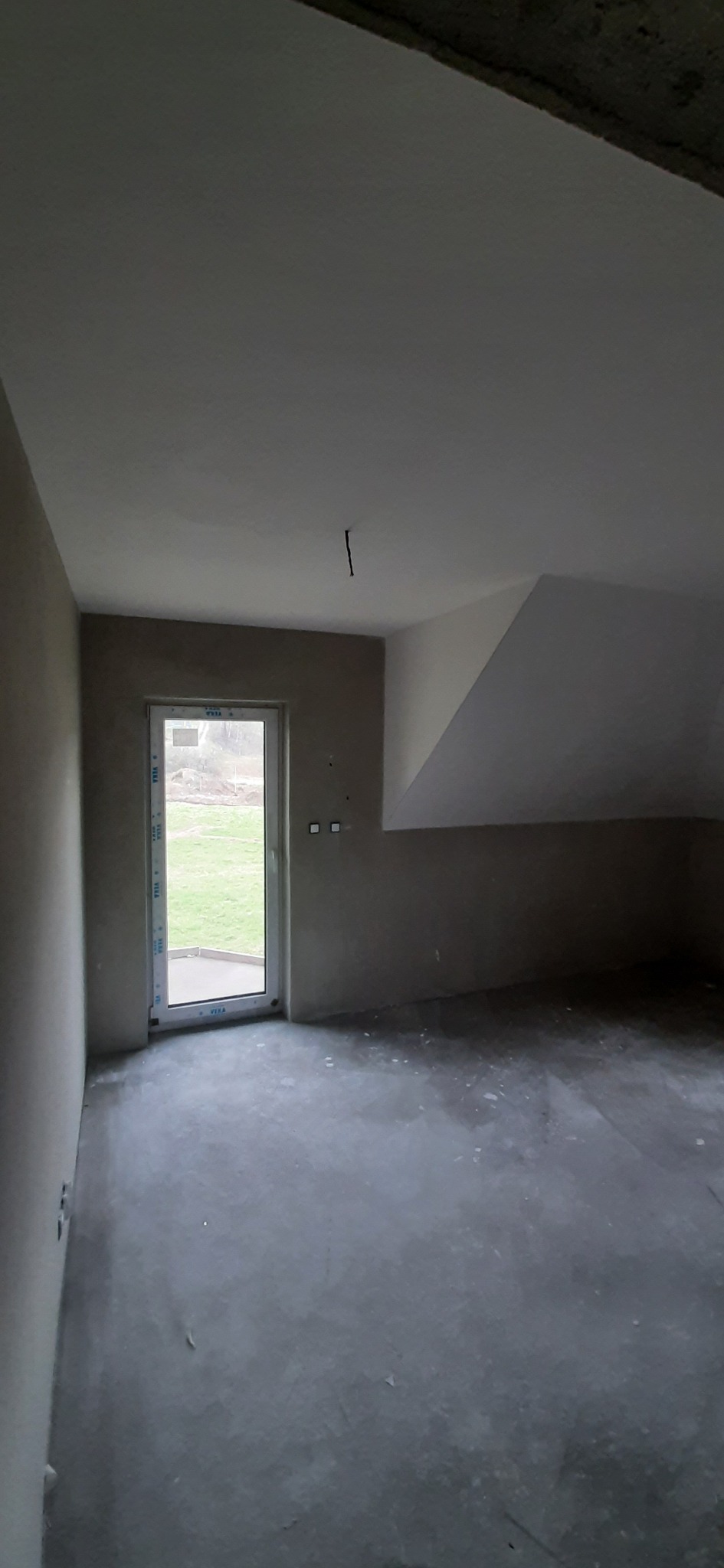 Surowe wnętrze poddasza z oknem balkonowym i widokiem na zieleń. Ściany przygotowane do malowania, sufit biały, podłoga betonowa. Widoczne gniazdka elektryczne.