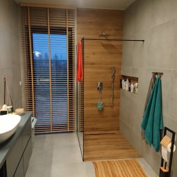 Renova - Nowoczesna łazienka z kabiną typu walk-in, drewnianymi akcentami i szarą armaturą. Widoczne rolety okienne i minimalistyczna umywalka nablatowa.