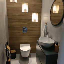 Renova - Nowoczesna łazienka w szarościach i drewnie. Umywalka nablatowa, wisząca toaleta, lustro w okrągłej ramie i drewniane wnęki z oświetleniem.