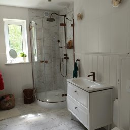 Renova - Jasna łazienka z kabiną prysznicową typu walk-in, białą szafką pod umywalką i marmurową podłogą. Armatura w kolorze miedzi. Dekoracyjne lampy na ścianie.