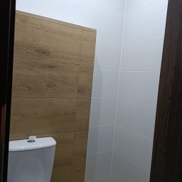 Renova - Nowoczesna toaleta po remoncie: białe płytki na ścianach kontrastują z drewnopodobnymi panelami. Widoczny fragment toalety i drzwi z ciemnego drewna.