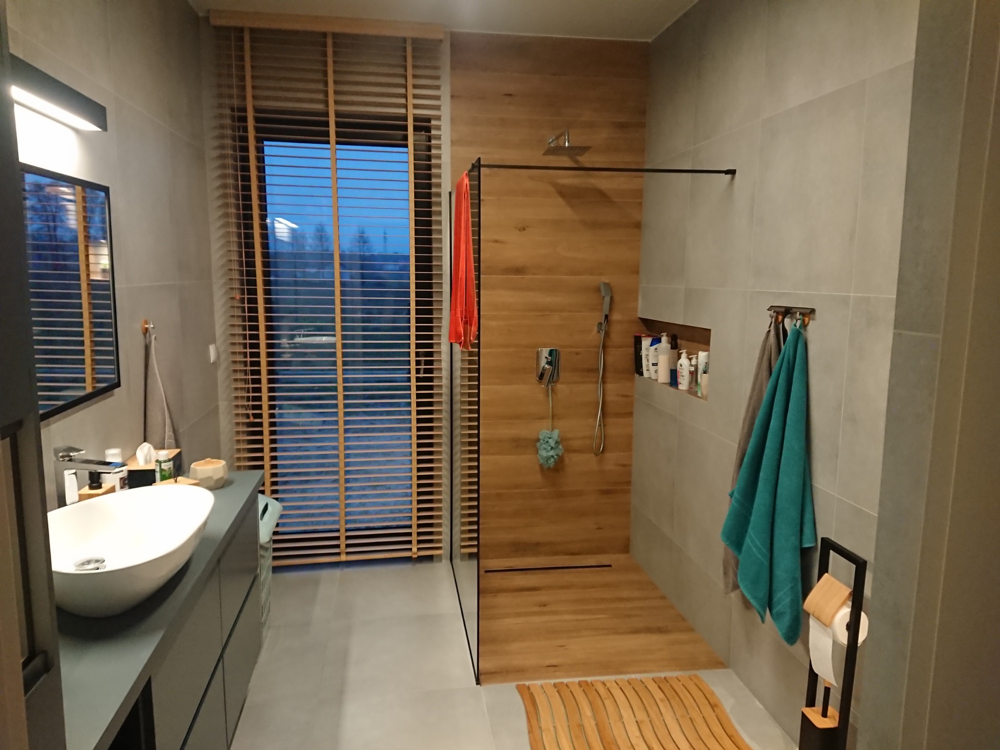 Nowoczesna łazienka z kabiną typu walk-in, drewnianymi akcentami i szarą armaturą. Widoczne rolety okienne i minimalistyczna umywalka nablatowa.