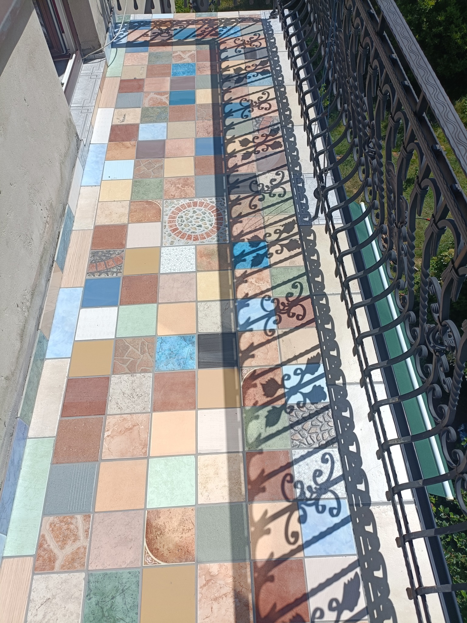 Kolorowa mozaika płytek na balkonie z kutą balustradą, rzucającą dekoracyjny cień. Nietypowe wzory i barwy płytek tworzą unikalny efekt wizualny.