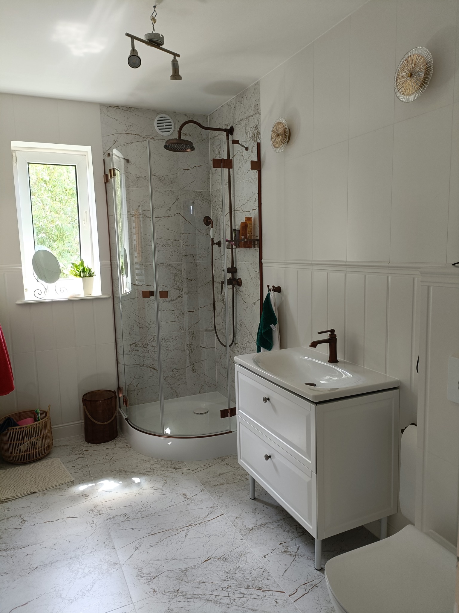 Jasna łazienka z kabiną prysznicową typu walk-in, białą szafką pod umywalką i marmurową podłogą. Armatura w kolorze miedzi. Dekoracyjne lampy na ścianie.