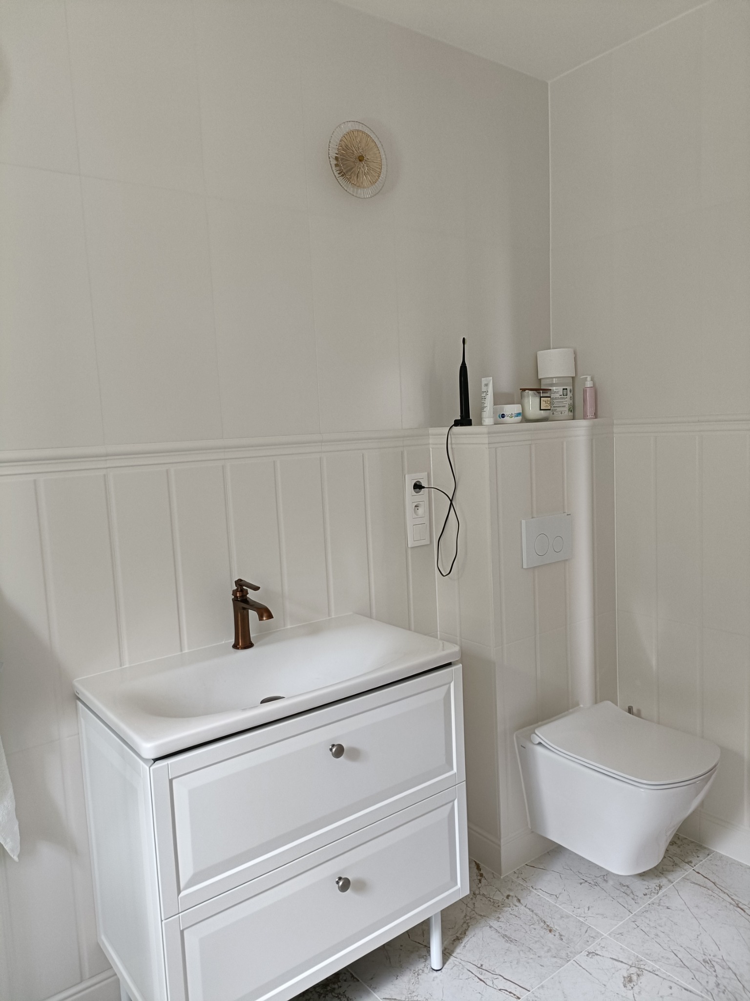 Jasna łazienka po remoncie: białe płytki, umywalka z szafką, toaleta podwieszana, marmurowe płytki podłogowe i dekoracyjny element na ścianie.