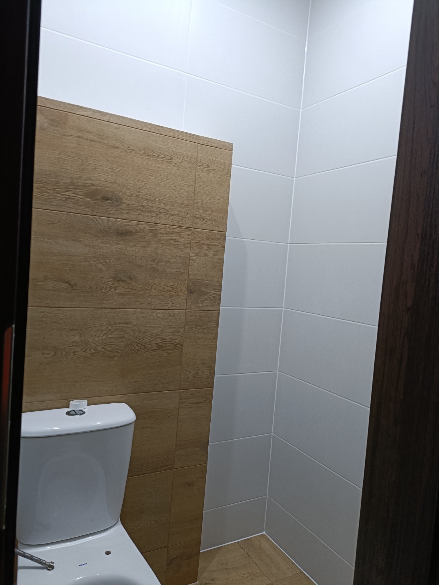 Nowoczesna toaleta po remoncie: białe płytki na ścianach kontrastują z drewnopodobnymi panelami. Widoczny fragment toalety i drzwi z ciemnego drewna.