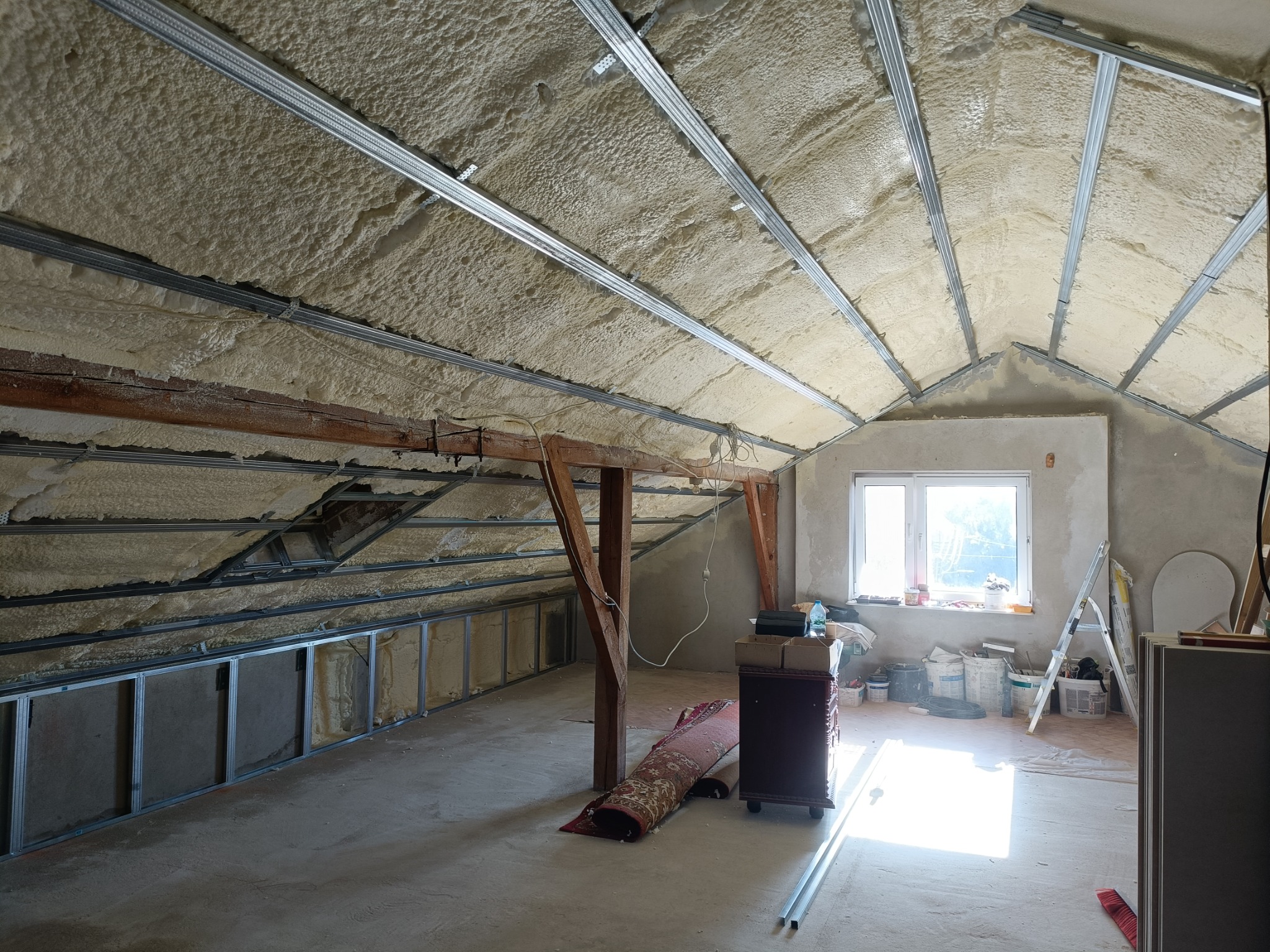 Poddasze po izolacji pianą PUR, widoczne metalowe profile pod płyty gipsowe. Fragment drewnianej konstrukcji i okno w tle. Częściowo zwinięty dywan i mebel na kółkach.