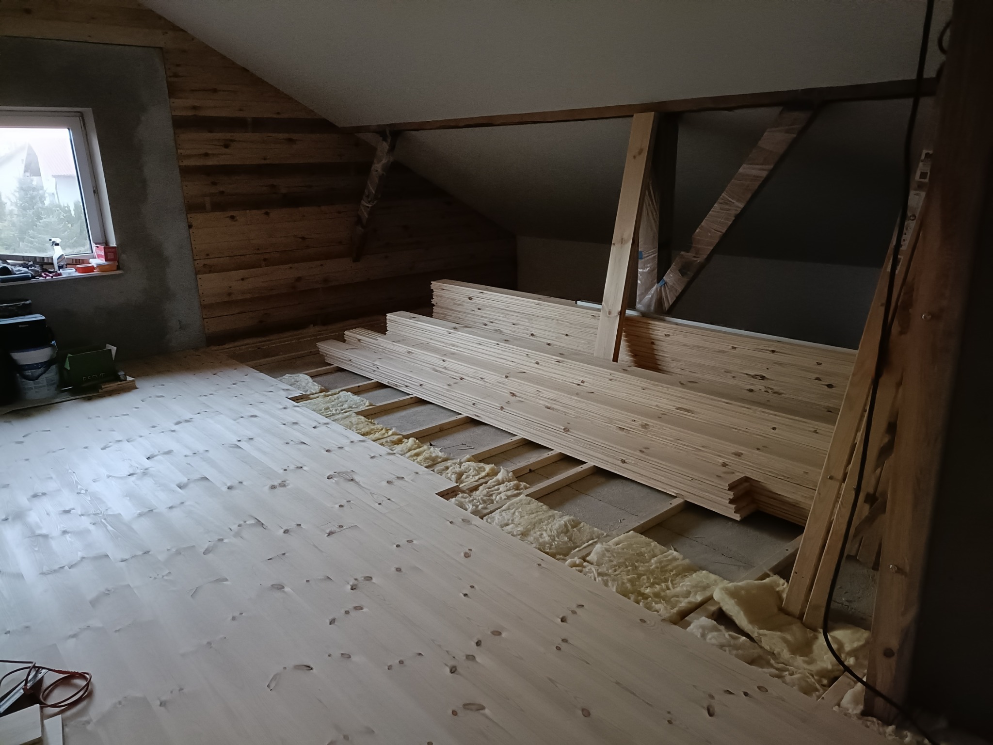 Podłoga z desek w trakcie montażu na poddaszu, widoczna izolacja. Częściowo wykończona drewniana ściana i okno. Ujęcie z góry, jasne drewno.