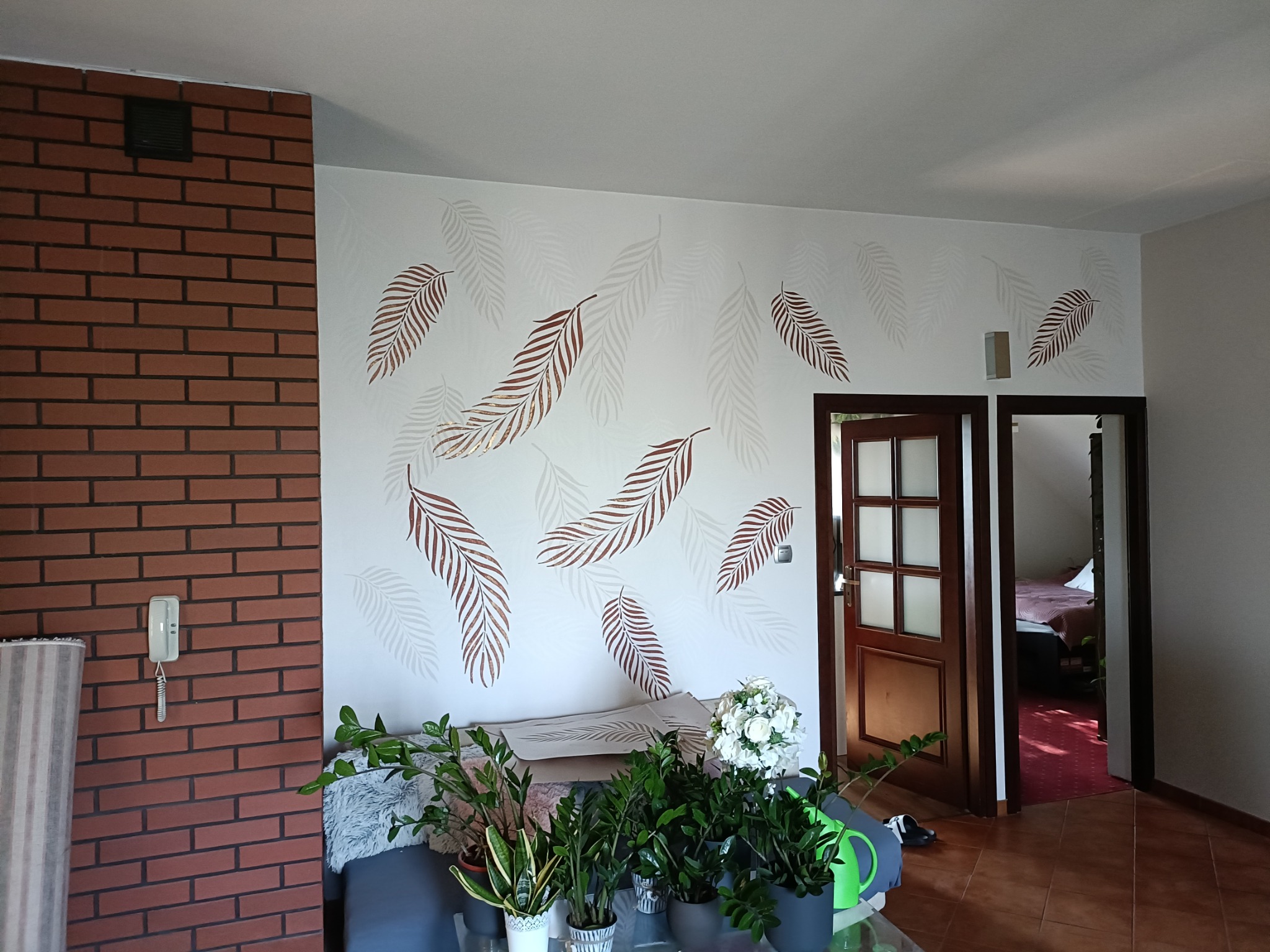 Pokój po malowaniu ścian z motywem liści, ceglana ściana z domofonem, rośliny doniczkowe na stole, otwarte drzwi do innego pomieszczenia.