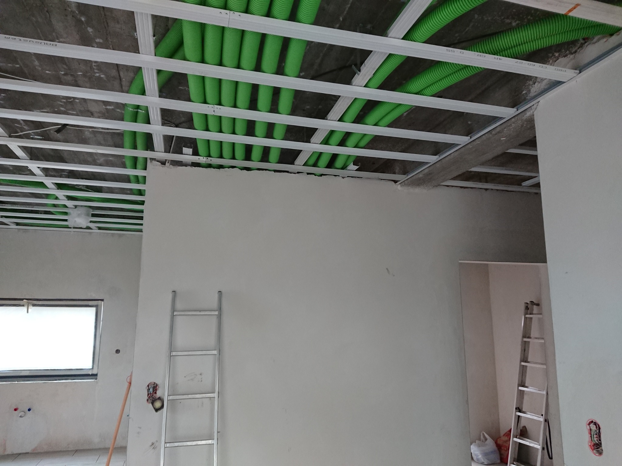 Sufit podwieszany w trakcie montażu z widocznymi zielonymi rurami instalacji elektrycznej. W tle ściany w surowym stanie i drabiny.
