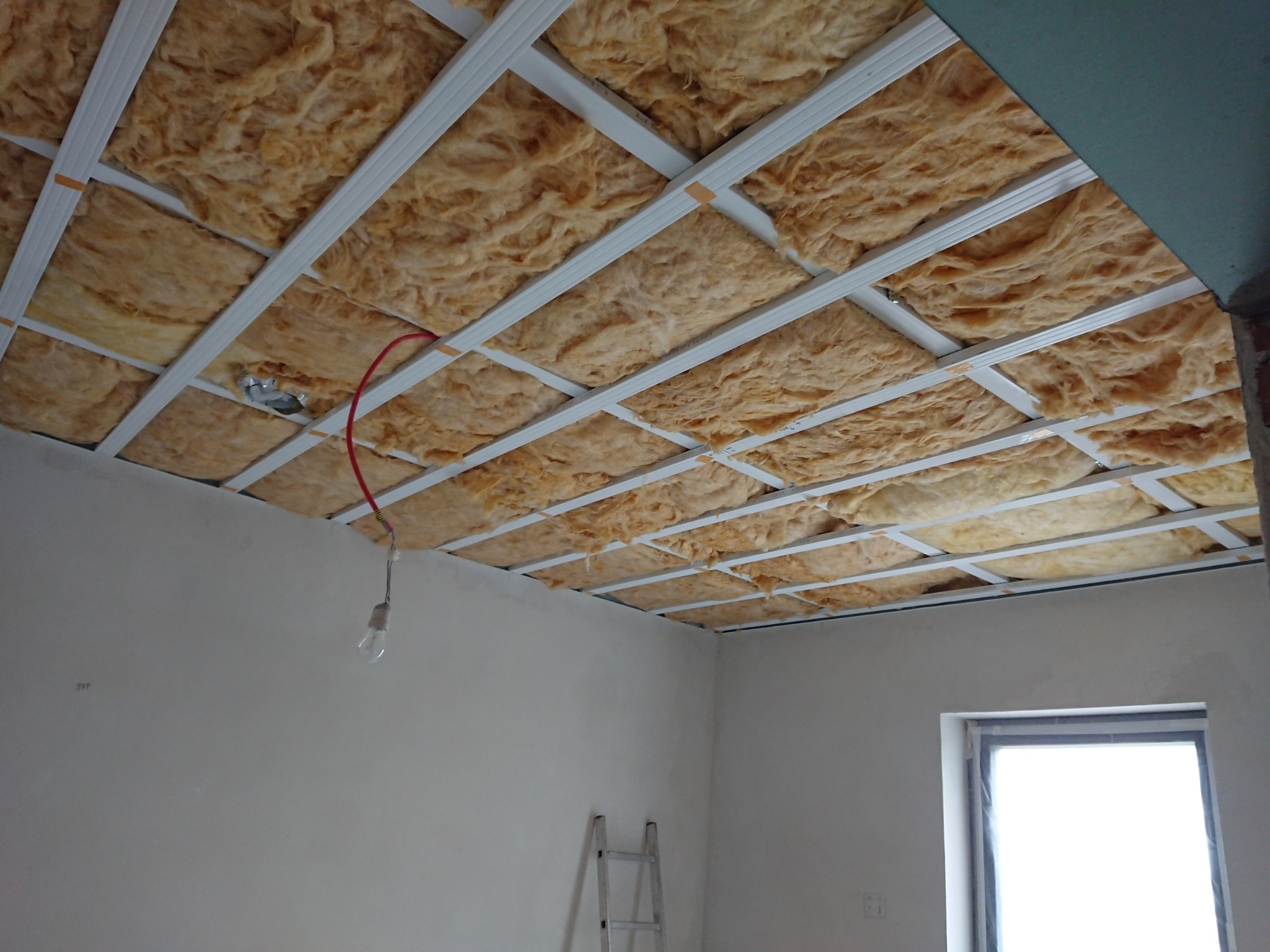 Montaż izolacji z wełny mineralnej na suficie podwieszanym z metalowym rusztem. Widoczna instalacja elektryczna i drabina. Etap prac wykończeniowych.