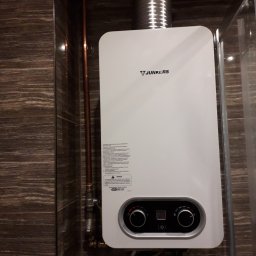 GP SMART ENERGY - Biały piec gazowy Junkers z podłączoną rurą odprowadzającą spaliny, zamontowany na ścianie wyłożonej brązowymi płytkami imitującymi drewno.