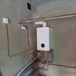 GP SMART ENERGY - Biały piec gazowy Bosch zamontowany na szarej ścianie z widocznymi rurami miedzianymi i izolowanymi, szarym tłem i elementami instalacji.
