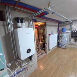 GP SMART ENERGY - Nowoczesny piec gazowy Bosch na ścianie, z widoczną instalacją grzewczą: rury miedziane i niebiesko-czerwone, grzejnik, zbiornik i czarne regulatory. Podłoga wyłożona panelami.