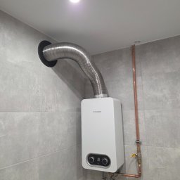 GP SMART ENERGY - Nowoczesny, biały piec gazowy Junkers z chromowaną rurą odprowadzającą spaliny, zamontowany na szarej, kafelkowanej ścianie w łazience. Widoczne miedziane rury i zawór.