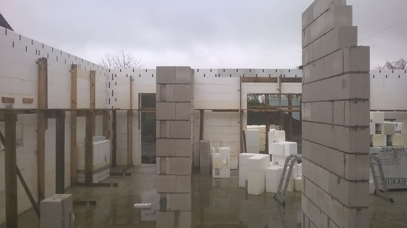 Budowa domu z bloczków w Gorzowie Wielkopolskim. Widoczne ściany z izolacją i drewniane podpory. Na podłodze woda, a w tle drzewa i pochmurne niebo.