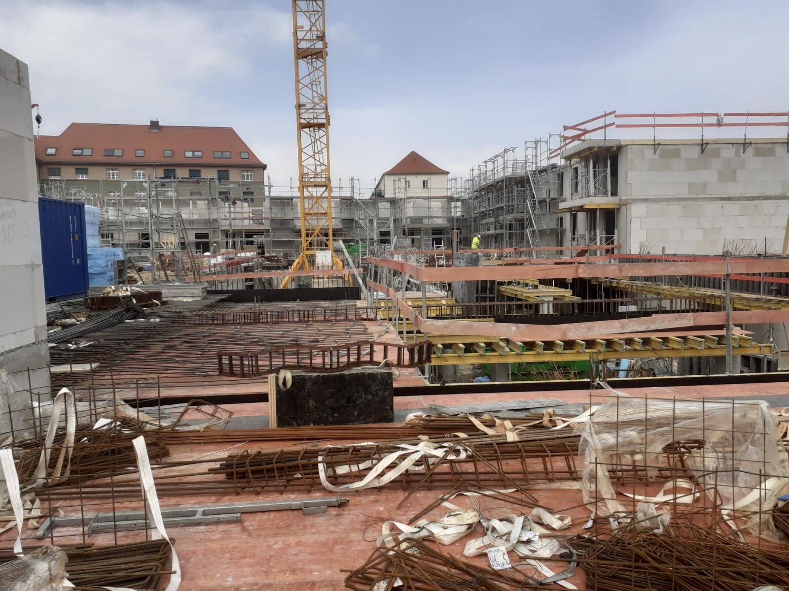 Budowa bloku mieszkalnego w toku: widok na zbrojenie stropu, dźwig i rusztowania. W tle budynki z czerwonymi dachami i pochmurne niebo.