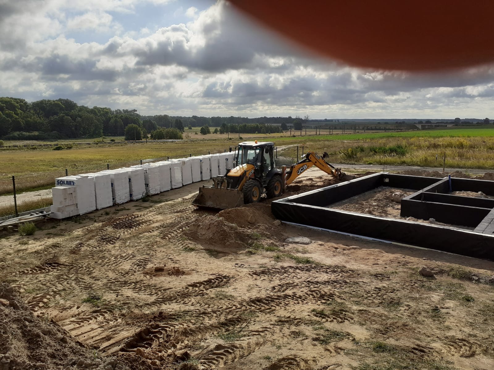 Fundamenty domu jednorodzinnego z hydroizolacją, obok palety z bloczkami Solbet i koparka JCB na placu budowy w słoneczny dzień. Widok z góry.