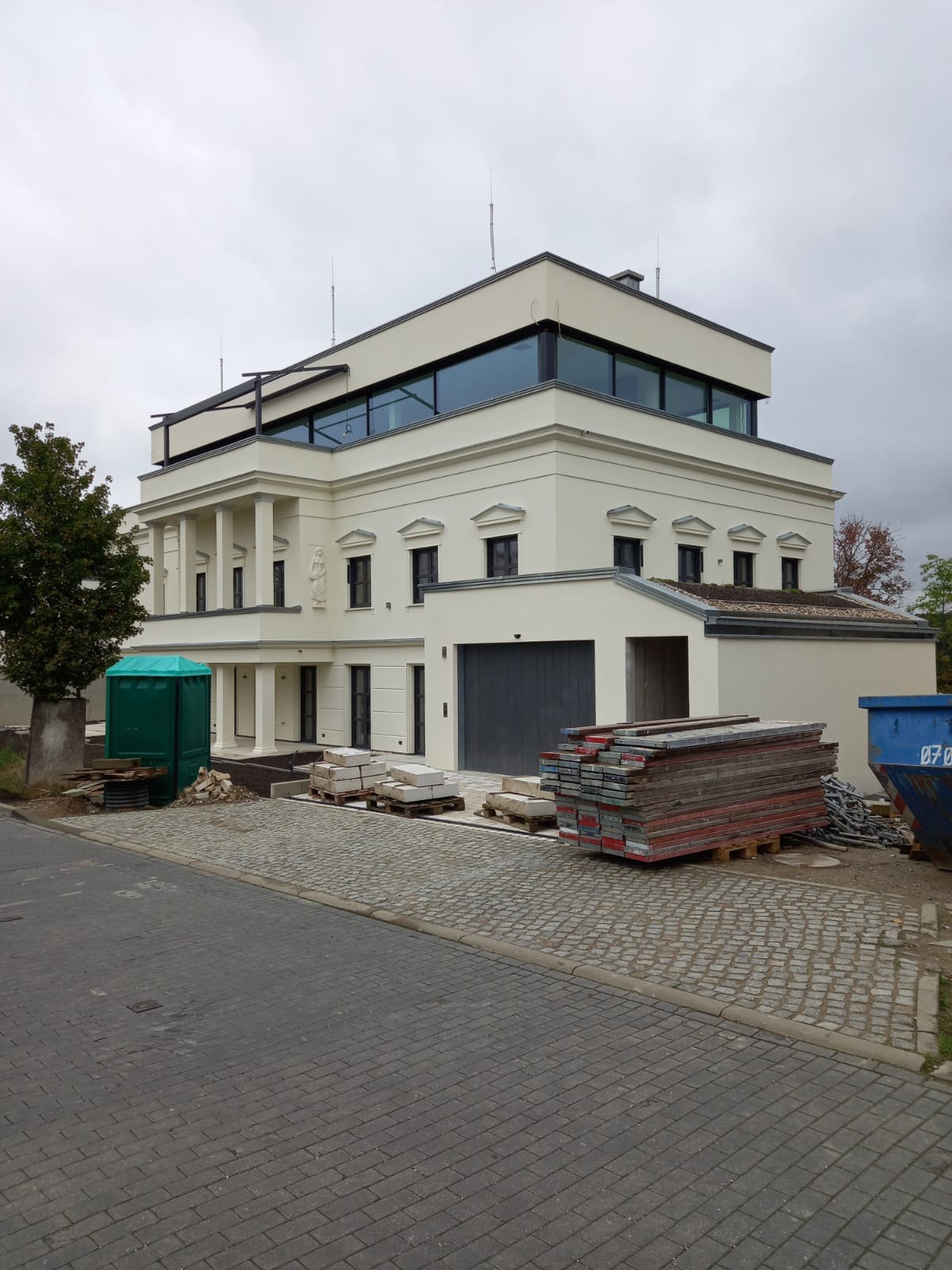 Elegancki, trzypiętrowy dom w stylu klasycystycznym z płaskim dachem i dużymi oknami, w trakcie budowy. Widoczne materiały budowlane i kontener na gruz.