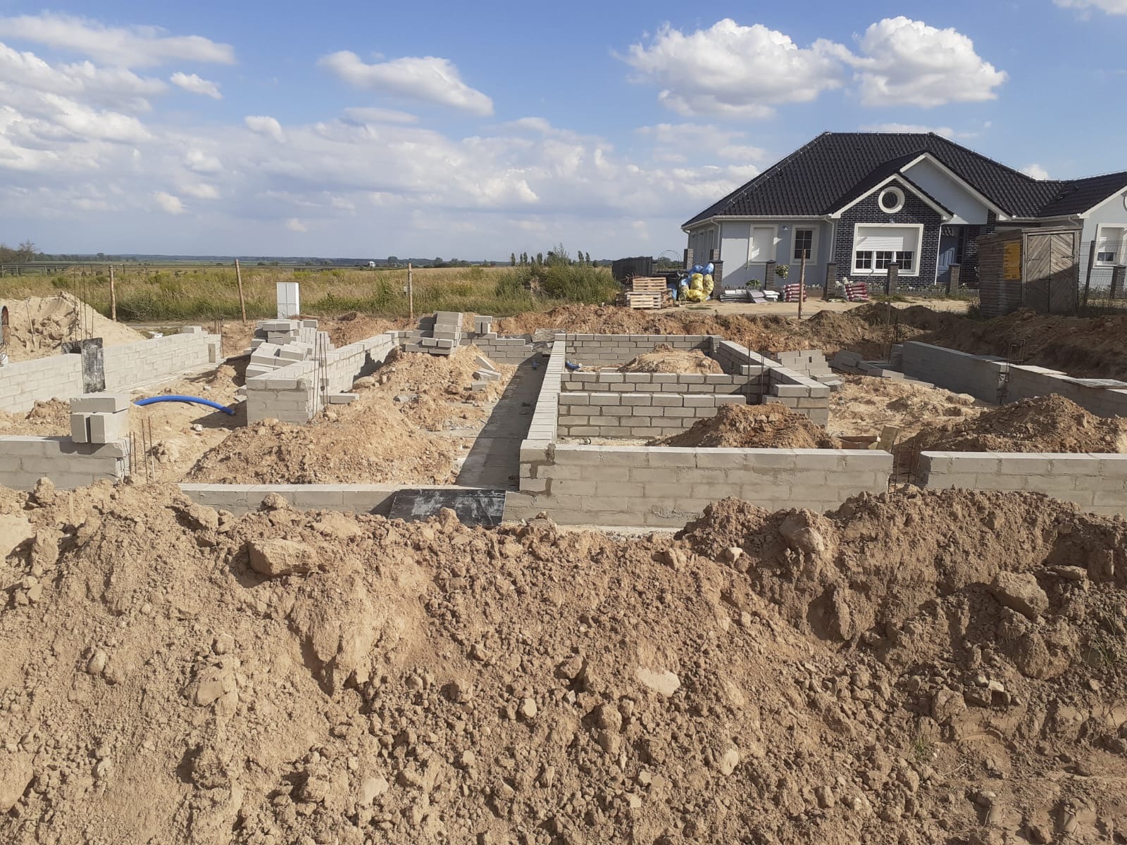 Fundamenty domu jednorodzinnego w trakcie budowy, widoczne bloczki betonowe i wykop, w tle dom w stanie surowym zamkniętym i błękitne niebo.
