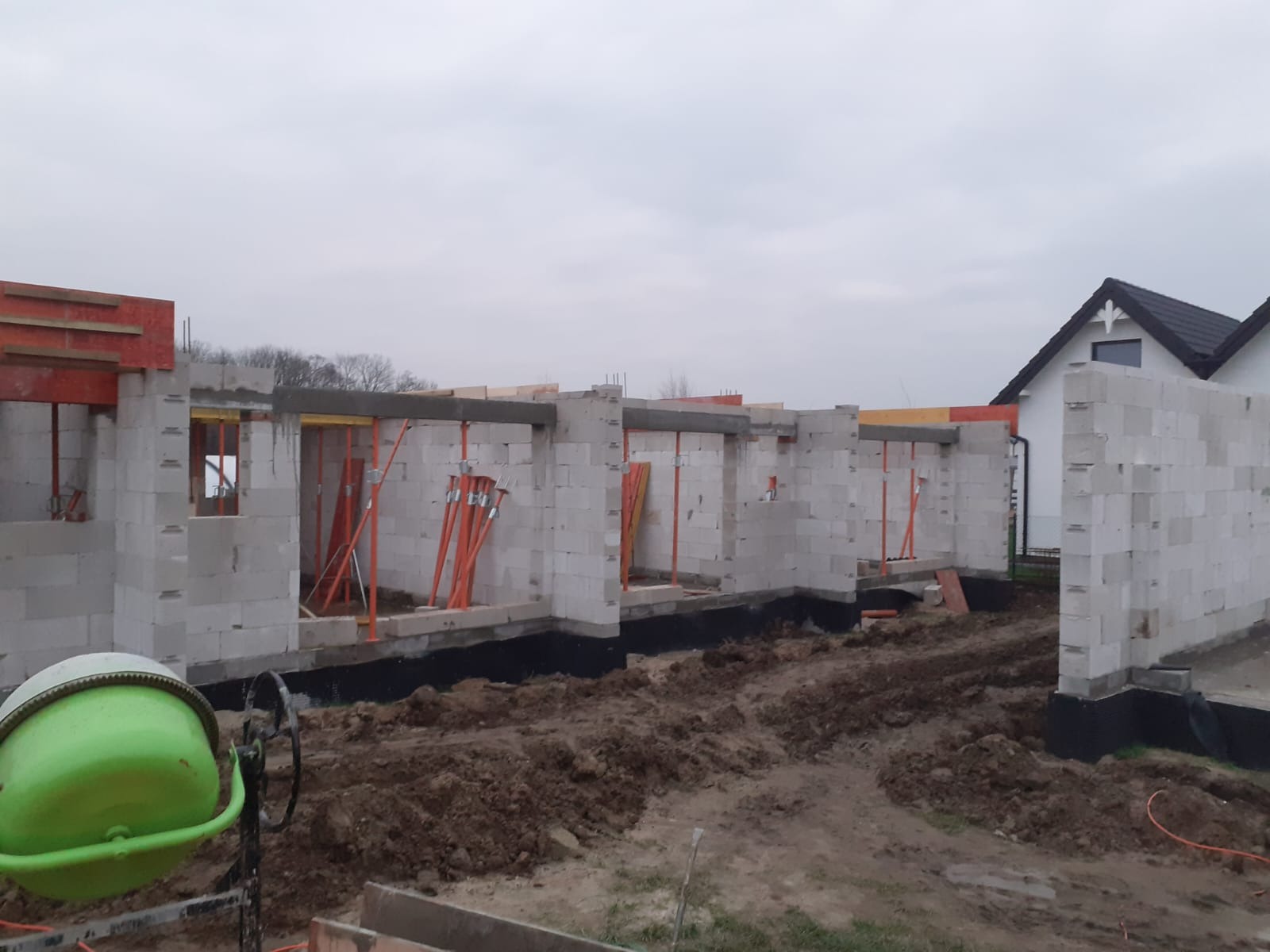Budowa domu w trakcie wznoszenia ścian z bloczków, widoczna betoniarka i podpory szalunkowe. Surowy stan, przygotowanie do dalszych prac.