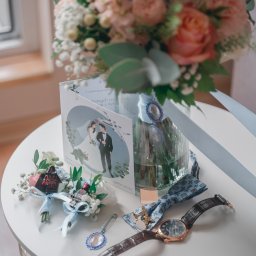 Pani-Migawka - Bukiet ślubny w wazonie, zaproszenie z parą młodą, mucha, zegarek i butonierki z motywem Star Wars na białym stoliku. Detale ślubne w pastelowych kolorach.