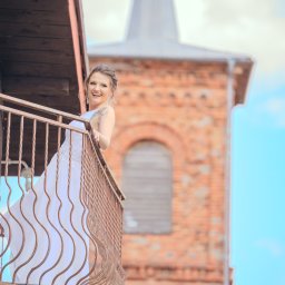 Pani-Migawka - Uśmiechnięta panna młoda w białej sukni na balkonie z metalową balustradą, w tle ceglany kościół z wieżą. Jasne, naturalne światło.