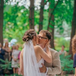 Pani-Migawka - Pocałunek pary młodej w plenerze, panna młoda w sukni z odkrytymi plecami i welonem, pan młody w beżowym garniturze i okularach, goście w tle, naturalne światło.